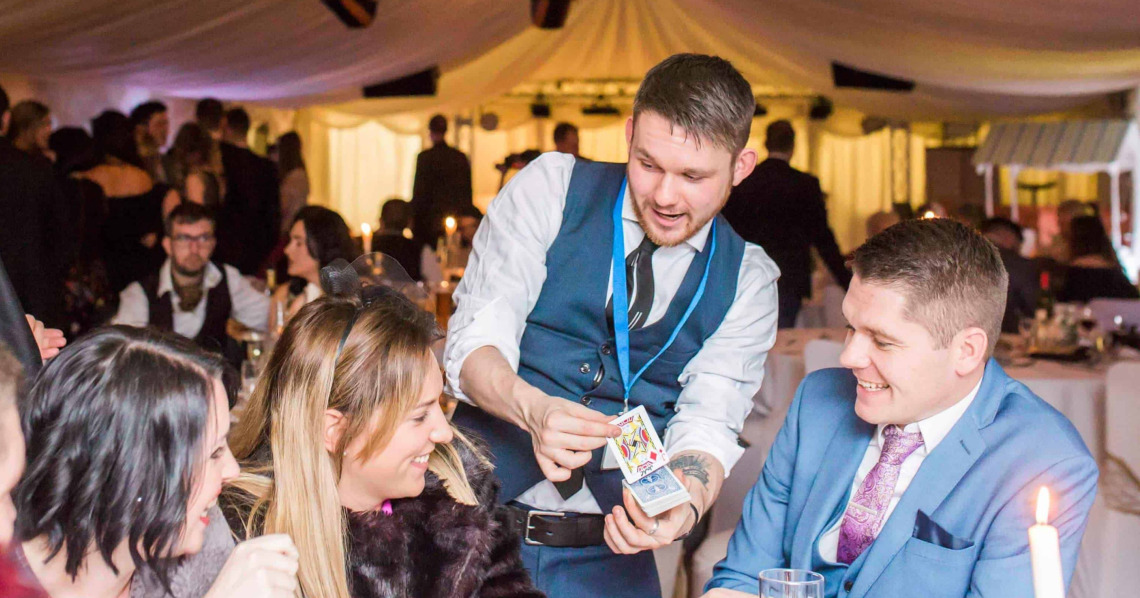 Greg Holroyd reveals a selected card during an evening wedding reception—guests burst out in laughter around the candlelit table.