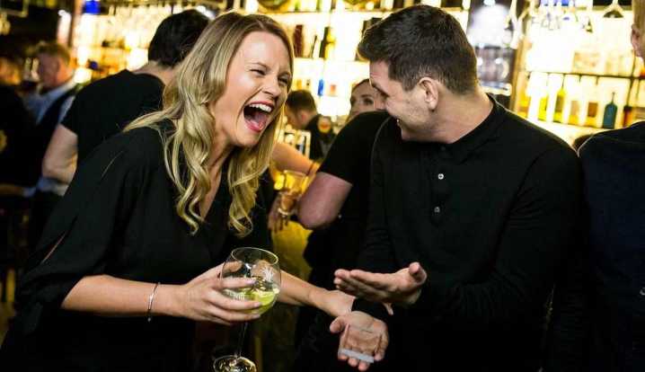 A woman bursts into laughter mid-drink as Greg Holroyd’s sleight of hand leaves the crowd speechless during a high-energy magic performance at a bar event.